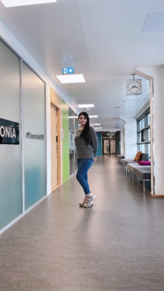 Nainen seisoo modernissa, hyvin valaistussa käytävässä, jossa on lasiseinät ja yläpuolella kello. Hänellä on harmaa villapaita, siniset farkut ja lenkkarit. "#SavoniaAMK" näkyy seinällä hänen vasemmalla puolellaan.