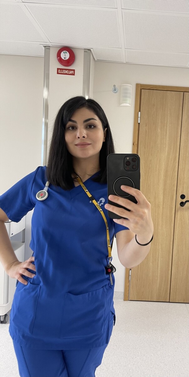 Siniseen lääkärinhoitohaalariin pukeutunut nainen seisoo hyvin valaistussa huoneessa ja ottaa peiliselfietä puhelimellaan. Hänellä on tummat hiukset, stetoskooppi ja kaulassaan keltainen kaulanauha. Hänen takanaan näkyy puinen ovi ja palovaroitin.