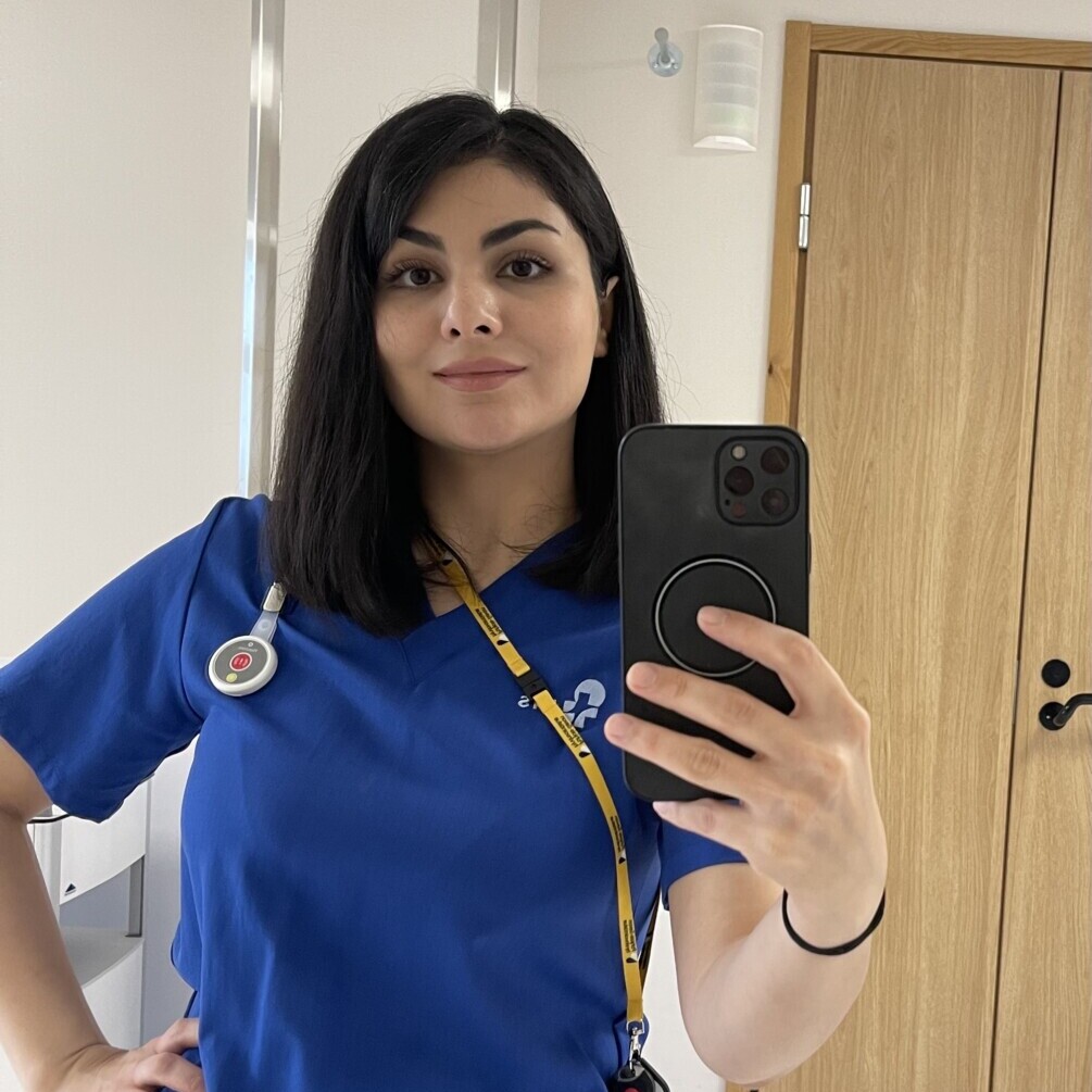 Siniseen lääkärinhoitohaalariin pukeutunut nainen seisoo hyvin valaistussa huoneessa ja ottaa peiliselfietä puhelimellaan. Hänellä on tummat hiukset, stetoskooppi ja kaulassaan keltainen kaulanauha. Hänen takanaan näkyy puinen ovi ja palovaroitin.