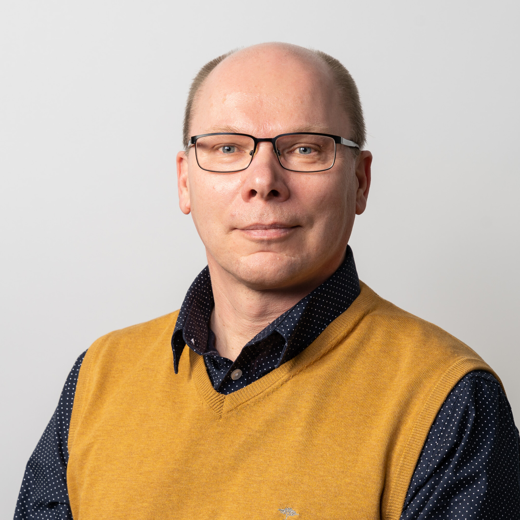 Keski-ikäinen mies, jolla on silmälasit, lyhyet vaaleanruskeat hiukset ja puhtaaksi ajetut kasvot, poseeraa vaaleaa taustaa vasten sinapinkeltaisessa villaliivissä ja tummansinisessä paidassa, jossa on valkoisia pisteitä.