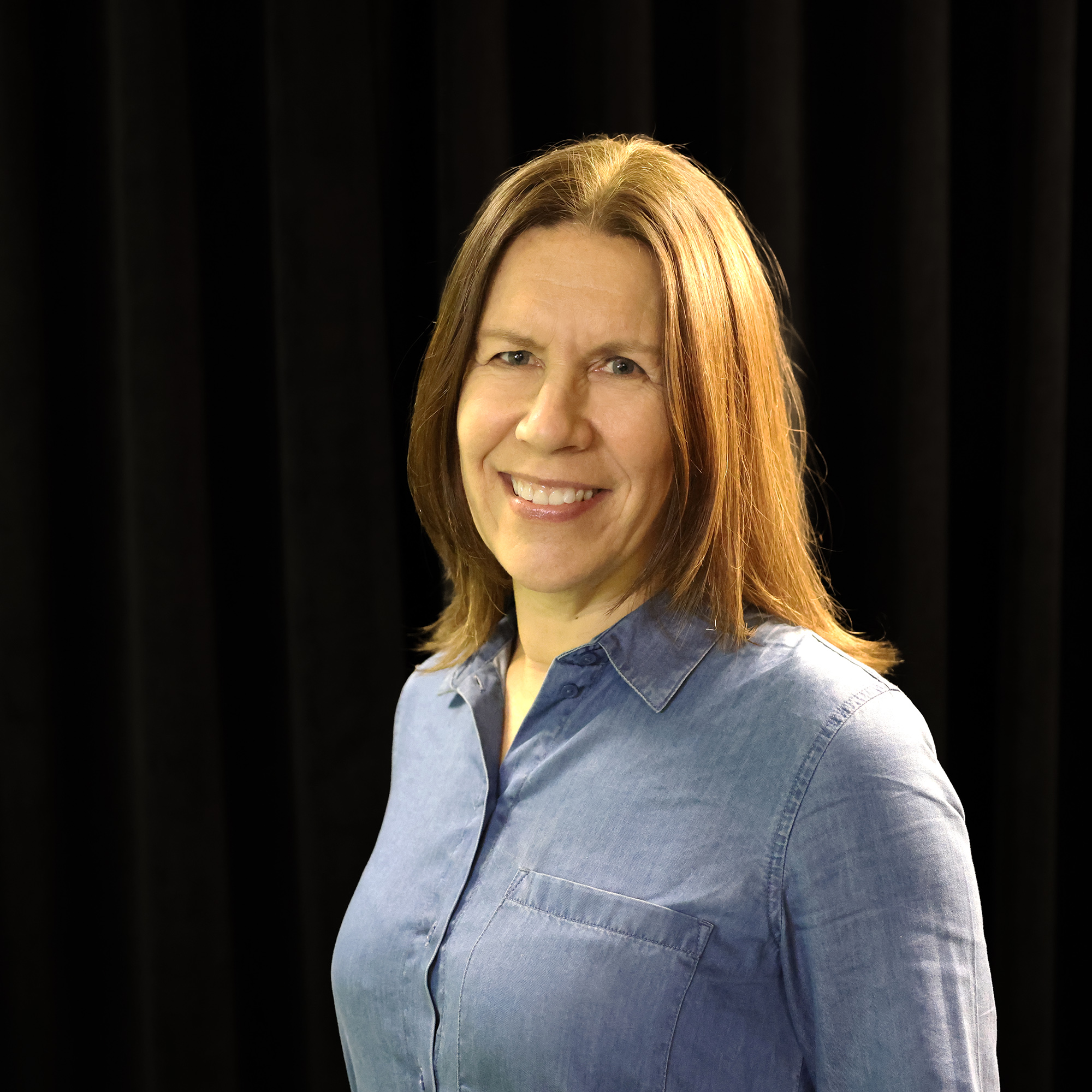 Nainen, jolla on suorat, hartioiden pituiset ruskeat hiukset, hymyilee seisoessaan mustan verhon edessä. Hänellä on yllään vaaleansininen napillinen paita.