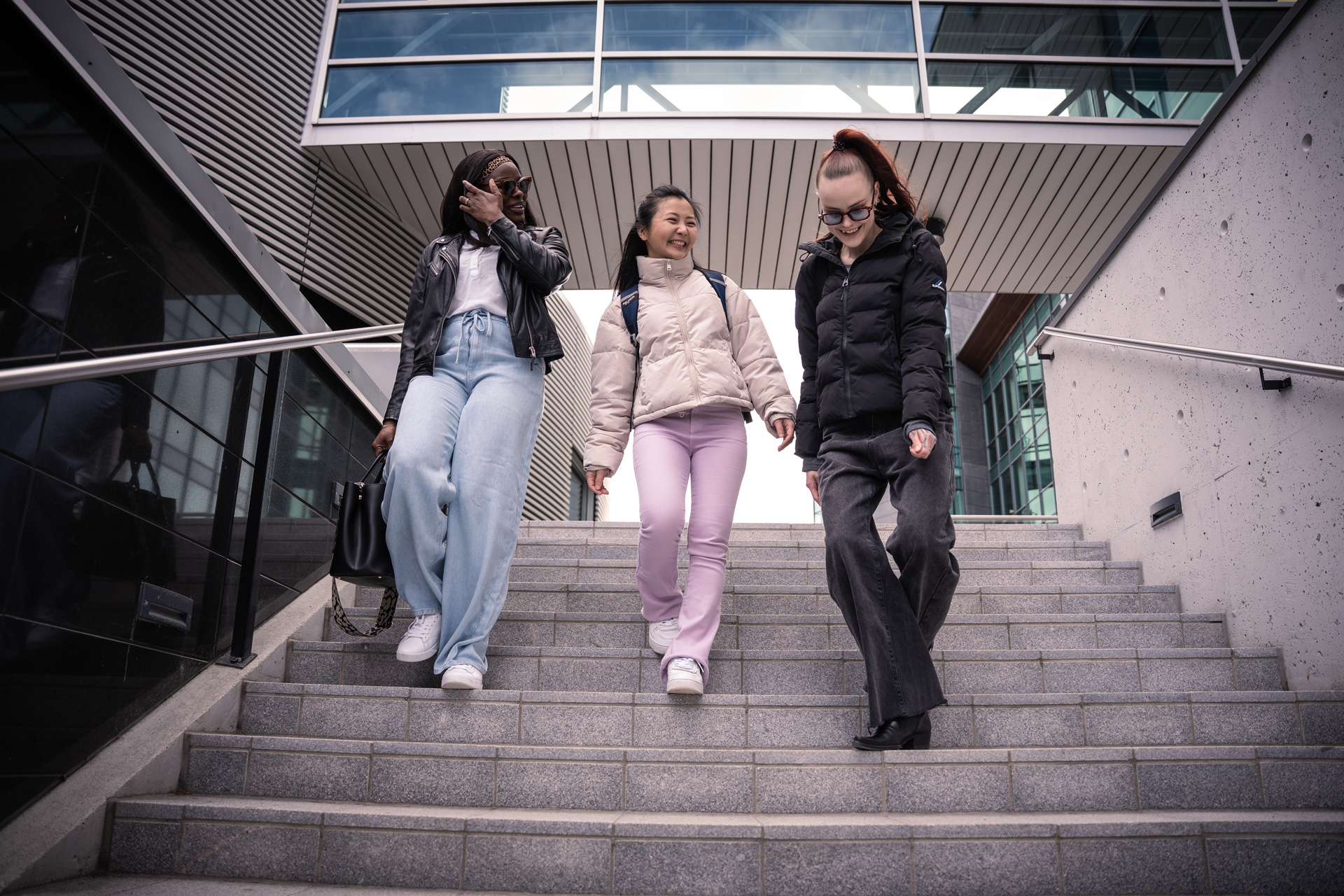 Kolme nuorta naista kävelee ulkoilmaportaita pitkin hymyillen ja keskustellen. Heillä on rennot talvivaatteet, takit ja farkut. Taustalla näkyy moderneja lasi- ja betonirakennuksia.