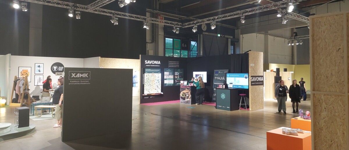 Tilava näyttelyhalli, jossa on erilaisia kojuja, näyttöjä ja tiedotustelineitä. Useita ihmisiä kävelee tai seisoo, ja alue on kirkkaasti valaistu ylävaloilla ja modernilla sisustuksella.