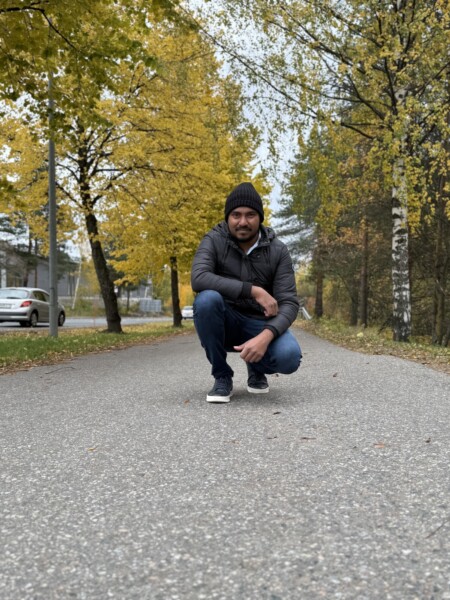 Mustaan pipoon, tummaan takkiin ja farkkuihin pukeutunut mies kyykistyy syksyisten puiden reunustamalla tyhjällä tiellä, jonka taustalla näkyy keltaisia ja vihreitä lehtiä ja pysäköityjä autoja.