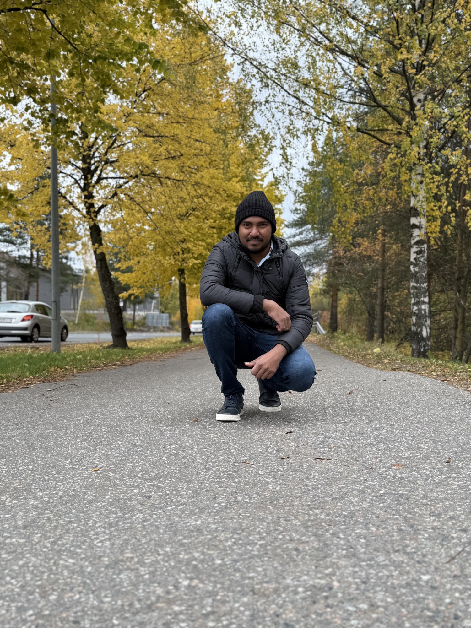 Mustaan pipoon, tummaan takkiin ja farkkuihin pukeutunut mies kyykistyy syksyisten puiden reunustamalla tyhjällä tiellä, jonka taustalla näkyy keltaisia ja vihreitä lehtiä ja pysäköityjä autoja.