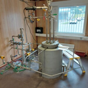 Suuri ruostumattomasta teräksestä valmistettu teollisuussäiliö, jossa on useita putkia, venttiilejä ja mittareita, on sijoitettu puuverhoiltuun huoneeseen, jossa on ikkuna, joka päästää päivänvalon sisään. Sähköjohtoja ja -laitteita on näkyvissä lattialla ja seinillä.