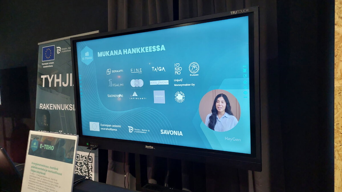 Suurella digitaalisella näytöllä näkyy sininen ruutu, jossa on suomenkielistä tekstiä, erilaisia yrityksen logoja ja naisen kuva. Näyttö on osa näyttelyosastoa, jonka lähellä on informatiivisia julisteita ja QR-koodi.
