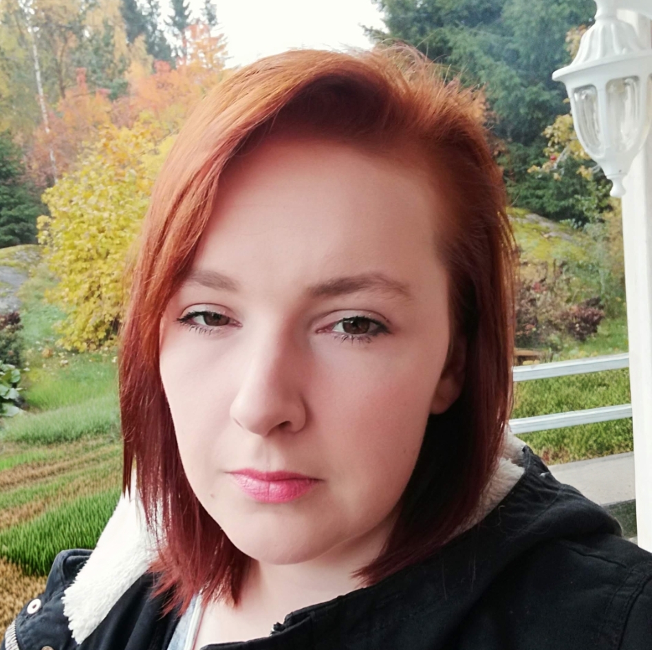 Ulkona seisoo nainen, jolla on suorat, hartioiden pituiset punaiset hiukset ja neutraali ilme. Hänellä on yllään musta takki, jossa on vaalea vuori. Taustalla näkyy puita ja syksyisiä lehtiä.