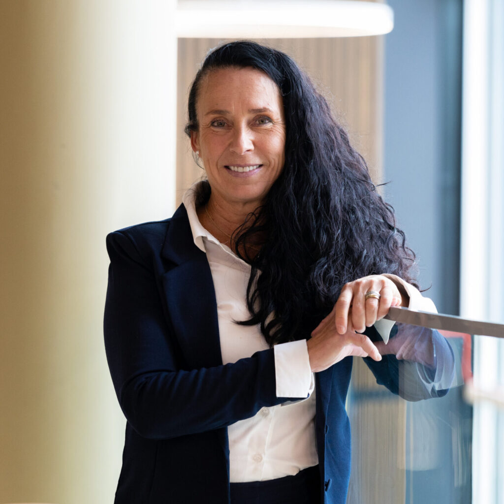 Nainen, jolla on pitkät, aaltoilevat tummat hiukset, laivastonsininen bleiseri ja valkoinen paita, seisoo sisätiloissa lasikaiteen vieressä ja hymyilee kameralle.