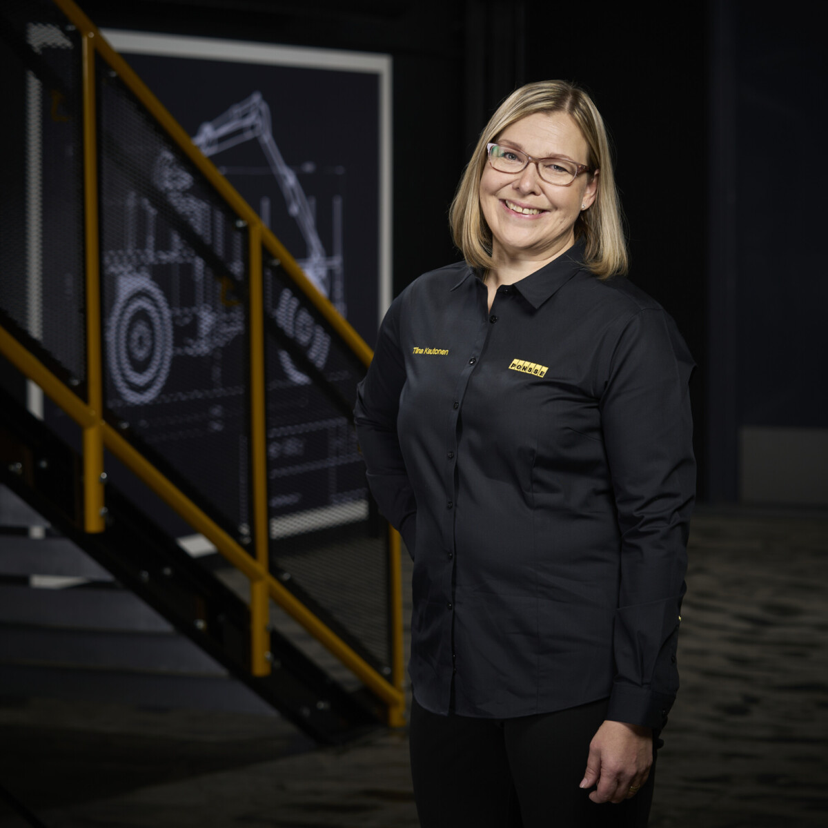 Sisällä seisoo hymyilevä vaaleatukkainen nainen, jolla on silmälasit ja musta napillinen paita, jossa on keltainen logo. Hänen takanaan on keltaisilla kaiteilla varustetut portaat ja suuri tekninen piirros rakennuskoneista.