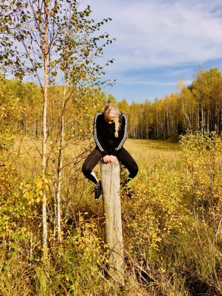 Vaaleahiuksinen, mustiin pukeutunut henkilö istuu puupylvään päällä syksyisellä pellolla, jota ympäröivät keltaiset puut sinisen taivaan alla.