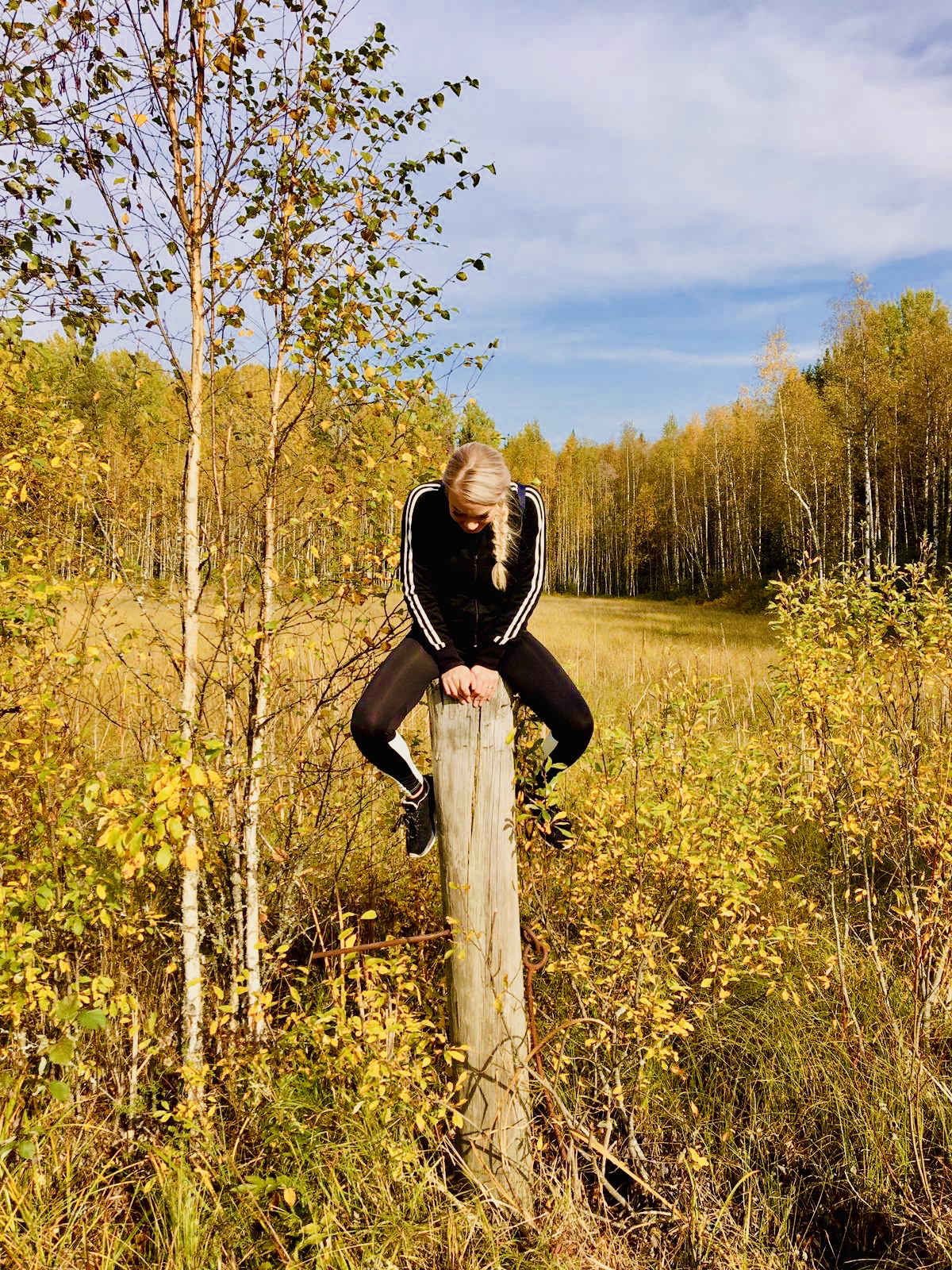 Vaaleahiuksinen, mustiin pukeutunut henkilö istuu puupylvään päällä syksyisellä pellolla, jota ympäröivät keltaiset puut sinisen taivaan alla.