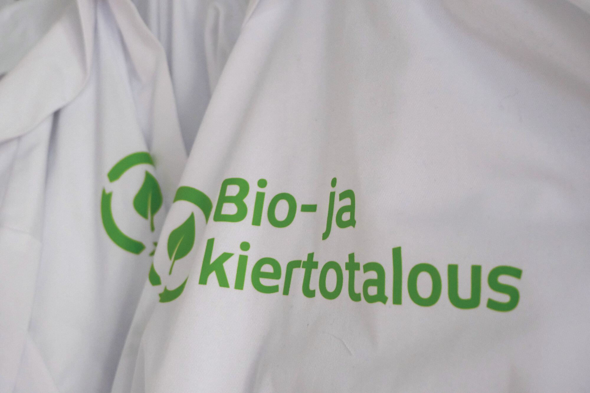 Valkoinen kangas, jossa on vihreä logo, jossa on kaksi ympyränuolia lehtien ympärillä ja suomenkielinen teksti Bio-ja kiertotalous, joka tarkoittaa bio- ja kiertotaloutta.