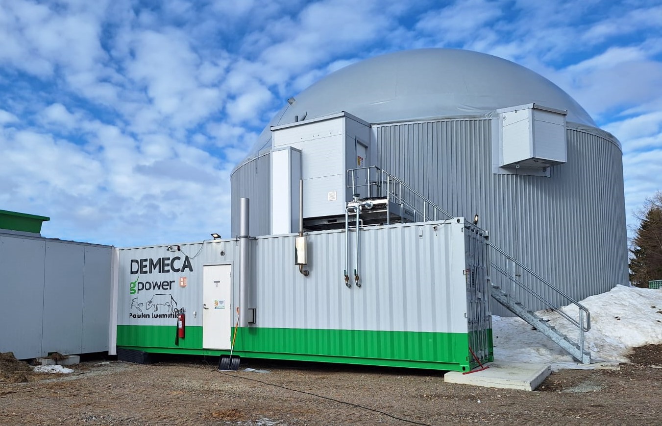 Suuri kupolimainen teollisuusrakennus, jonka ulkopinta on aaltopahvista terästä, seisoo vihreän ja valkoisen kontin takana, jossa lukee "DEMECA gpower". Oviin johtavat portaat, ja maassa on lunta osittain pilvisen taivaan alla.