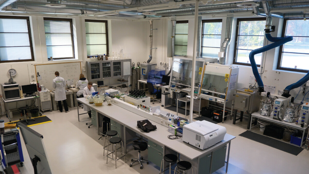 Nykyaikainen laboratorio, jossa kaksi laboratoriotakkiin pukeutunutta ihmistä työskentelee työpisteiden äärellä, ja ympärillä on laitteita, tietokoneita, lasitavaroita ja ilmanvaihtojärjestelmiä. Suuret ikkunat päästävät luonnonvaloa sisään. Tila vaikuttaa siistiltä ja järjestetyltä.