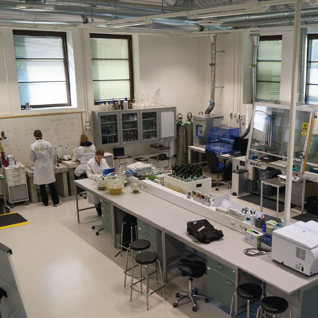 Tilava laboratorio, jossa kaksi valkoisiin laboratoriotakkeihin pukeutunutta ihmistä työskentelee eri työpisteissä. Huoneessa on laboratoriolaitteita, tietokoneita, lasitavaroita ja kaappeja, ja suuret ikkunat päästävät luonnonvaloa sisään.