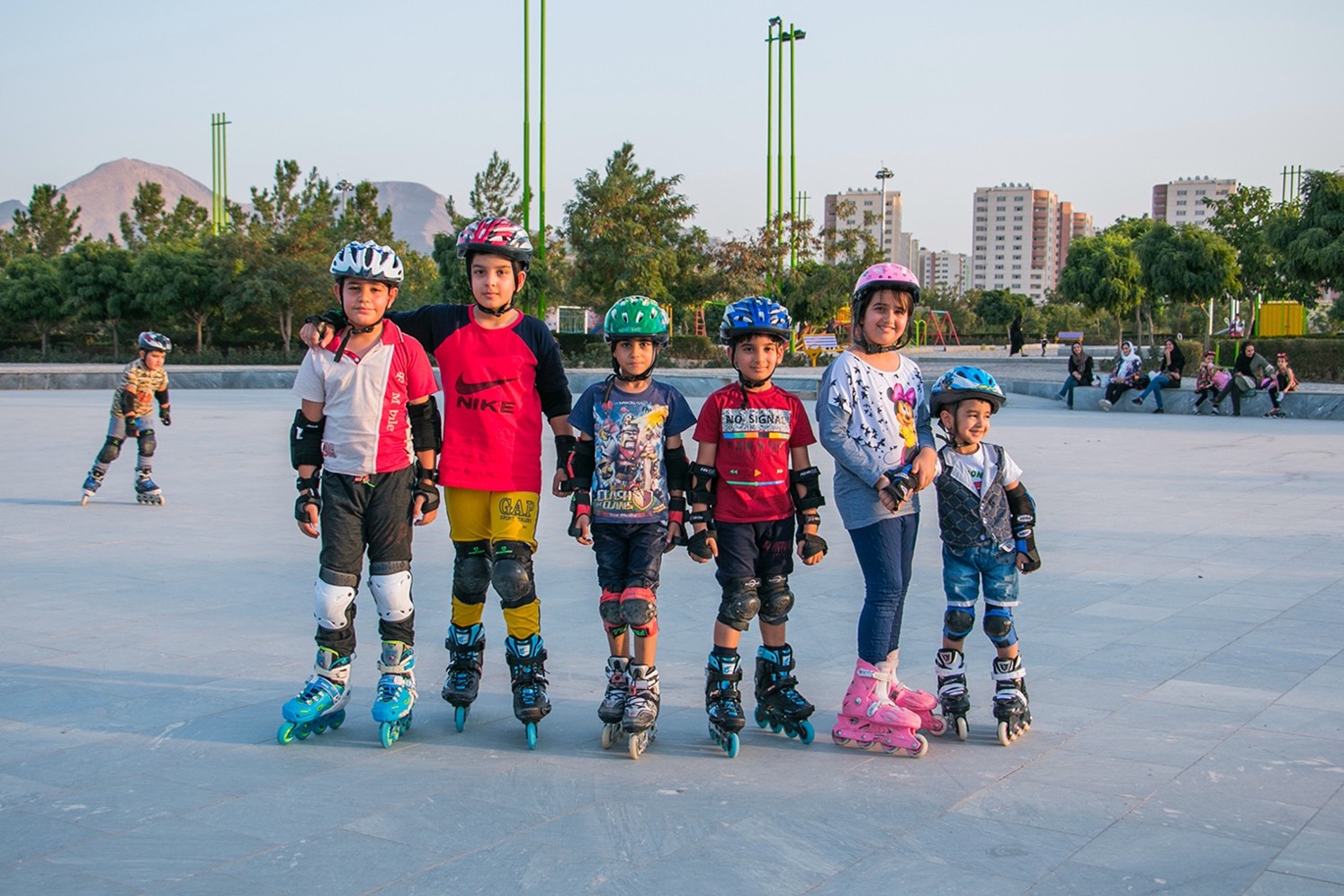 Kuusi lasta, joilla on kypärät ja rullaluistimet, seisoo rivissä ulkona luistelualueella ja hymyilee kameralle. Taustalla näkyy rakennuksia, puita ja muita ihmisiä.