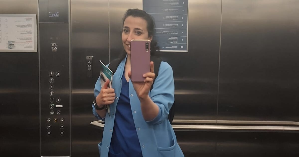 Siniseen lääkärinhoitohaalariin ja vaaleansiniseen takkiin pukeutunut nainen seisoo hississä ja pitää puhelinta kädessään ottaakseen peiliselfien. Hän hymyilee, pitää papereita kädessään ja näyttää peukkua ylöspäin. Hissin painikkeet ja metalliseinät ovat näkyvissä.