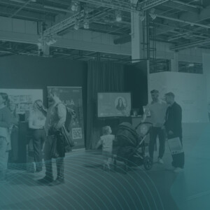 Ihmiset seisovat ja ovat vuorovaikutuksessa sisätiloissa pidettävissä näyttelyissä tai konferensseissa, joissa on esittelykoppeja, näyttöjä ja banderolleja. Kuvassa on tiilenpunainen päällys ja etualalla abstrakti viivakuvio.