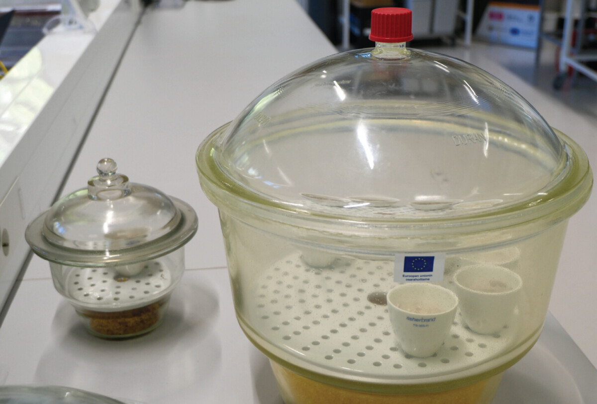 Laboratoriossa on kaksi lasista eksikaattoria. Suuremmassa on useita pieniä valkoisia laboratorioaltaita, kun taas pienemmässä on rakeista ainetta. Molemmissa on kirkkaat kannet, toisessa punainen nuppi. Taustalla näkyy laboratoriolaitteita.