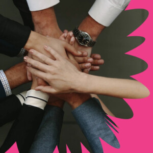 Useita eri ihmisten käsiä on pinottu yhteen tiimityön eleenä, ja taustalla on kirkkaan vaaleanpunainen abstrakti muoto.