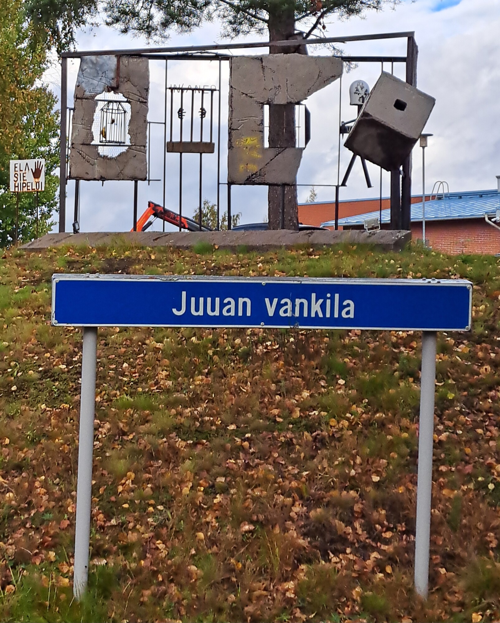 Sinisessä kyltissä, jossa on valkoinen teksti, lukee Juuan vankila. Sen takana nurmikumpareella on rikkinäistä betonivankilan seinää muistuttava taideinstallaatio, jossa on kaltereita ja esineitä. Taustalla on puita ja rakennus.