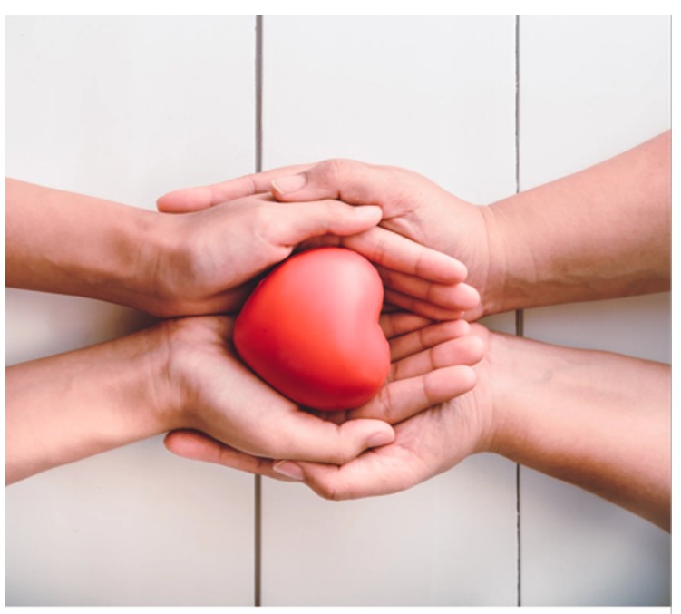 Kaksi käsiparia pitelee hellästi punaisen sydämen muotoa valkoisella puupinnalla, mikä symboloi huolenpitoa, rakkautta ja tukea.