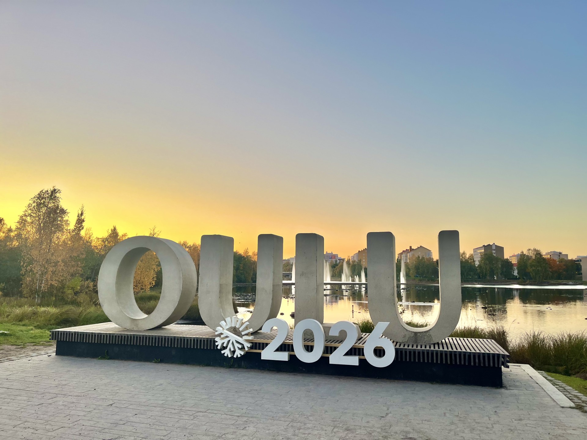 Suuret valkoiset kirjaimet, joissa lukee OULU 2026, seisovat järven rannalla auringonlaskun aikaan, ja lumihiutaleen symboli on 2026. Taustalla näkyy puita ja rakennuksia, jotka heijastuvat tyynestä vedestä.