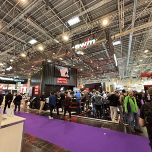 Kiireinen messutapahtuma, jossa monet ihmiset kävelevät ja kokoontuvat suuren EWM:n ständin lähellä teollisuusvalojen alla; punainen Welding in Action -kyltti on näkyvissä, ja etualalla on violetti matto.