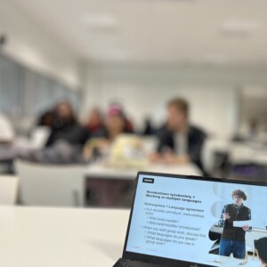 Sumea luokkahuone, jossa oppilaat istuvat pöydissä, avoimen kannettavan tietokoneen takaa kuvattuna. Kannettavan tietokoneen näytöllä näkyy esitysdia monikielisestä työskentelystä, jossa on tekstiä ja kuva puhuvasta henkilöstä.