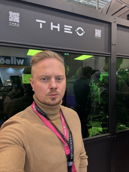 Mies, jolla on lyhyet vaaleat hiukset ja leikattu parta, ottaa selfien mustan, vihreällä valaistulla THEO-näytön edessä. Miehellä on ruskea poolopusero ja vaaleanpunainen kaulanauha. Taustalla näkyy ihmisten heijastuksia.