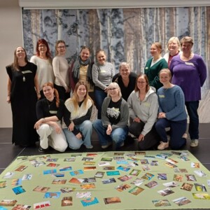 Viidentoista hymyilevän ihmisen ryhmä poseeraa sisätiloissa mustalla lattialla, jossa on vihreä matto, joka on peitetty erilaisilla korteilla. Heidän takanaan on seinä, jossa on koivumetsämaalausta ja sininen banderolli, jossa lukee: "Potential unlimited".