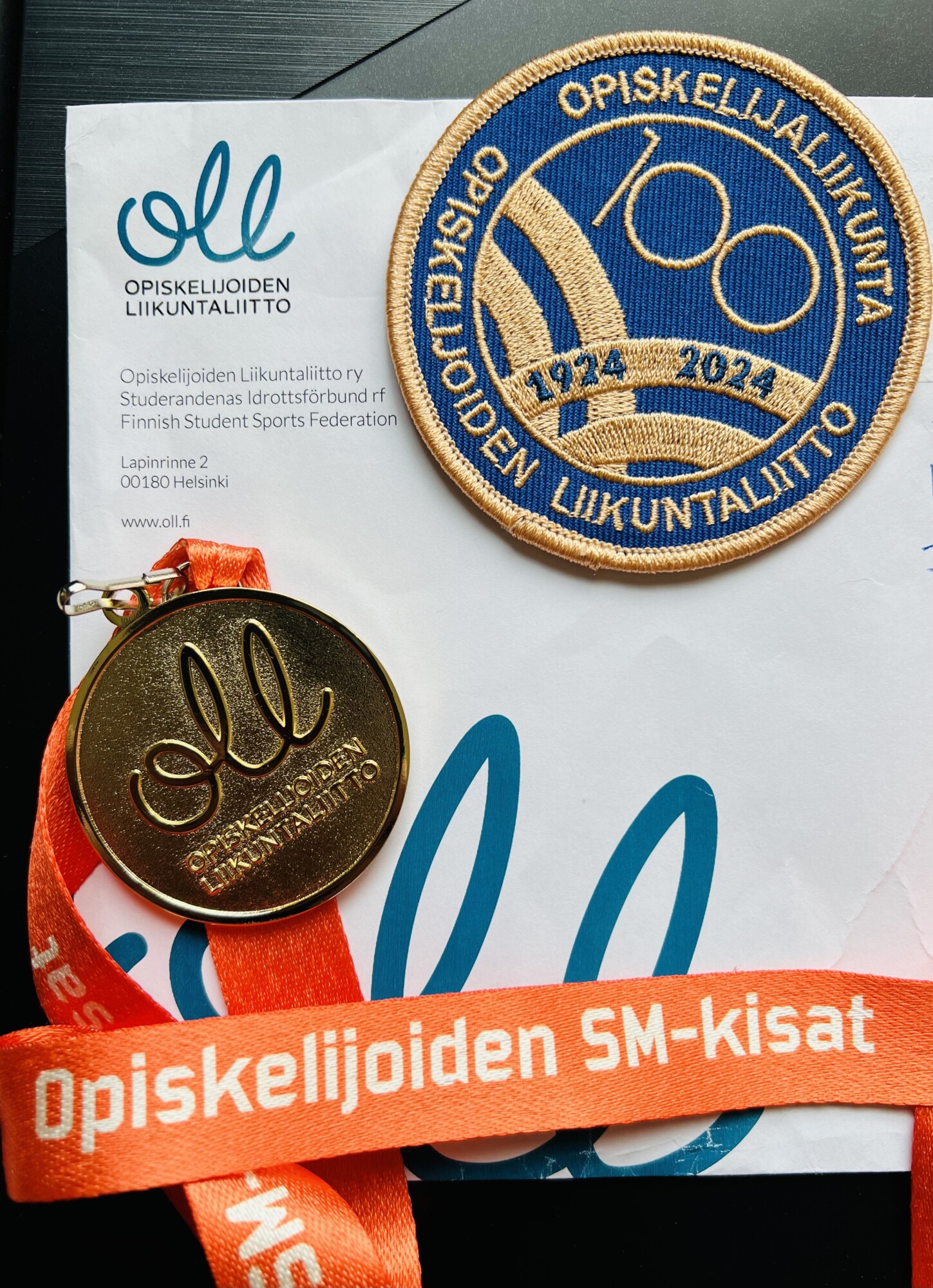 Mitali, jossa on oranssi kaulanauha, pyöreä muistolaastari ja Suomen Ylioppilasurheiluliiton (OLL) kirje, jossa juhlistetaan 100-vuotista taivalta (1924-2024) ja Opiskelijoiden SM-kisat -urheilutapahtumaa.