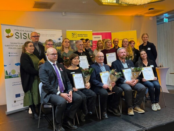 Joukko ihmisiä, joista osa istuu ja osa seisoo, poseeraa sisätiloissa pitelemässä kehystettyjä todistuksia ja kukkakimppuja. Taustalla näkyy bannereita, joissa on logoja ja suomenkielistä tekstiä.