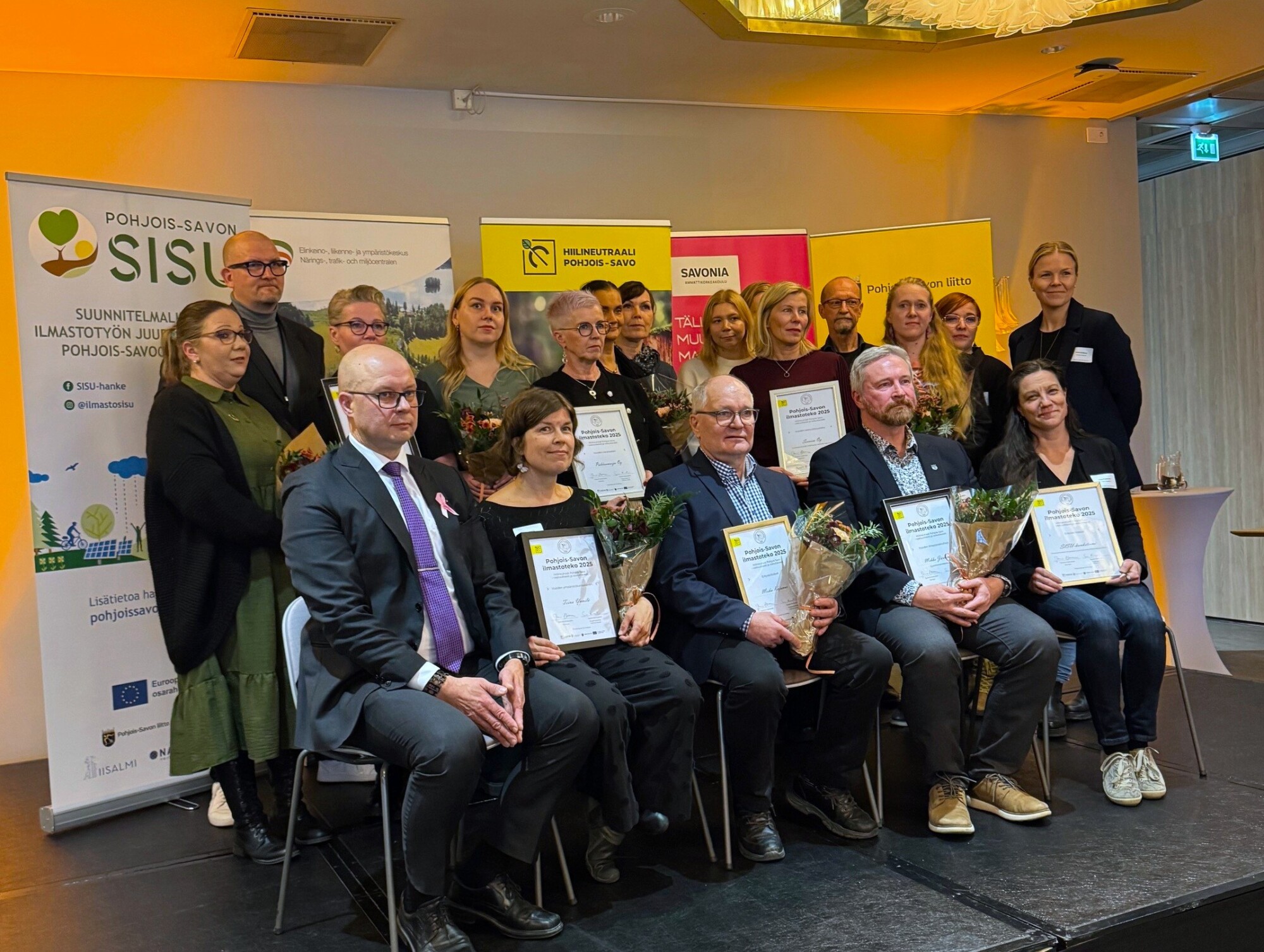 Joukko ihmisiä, joista osa istuu ja osa seisoo, poseeraa sisätiloissa pitelemässä kehystettyjä todistuksia ja kukkakimppuja. Taustalla näkyy bannereita, joissa on logoja ja suomenkielistä tekstiä.