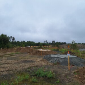 Avoin, hiekkainen työmaa, jossa on useita puisia merkkejä ja pieniä sorakasoja. Puut ja pensaat reunustavat aluetta pilvisen, pilvisen taivaan alla.