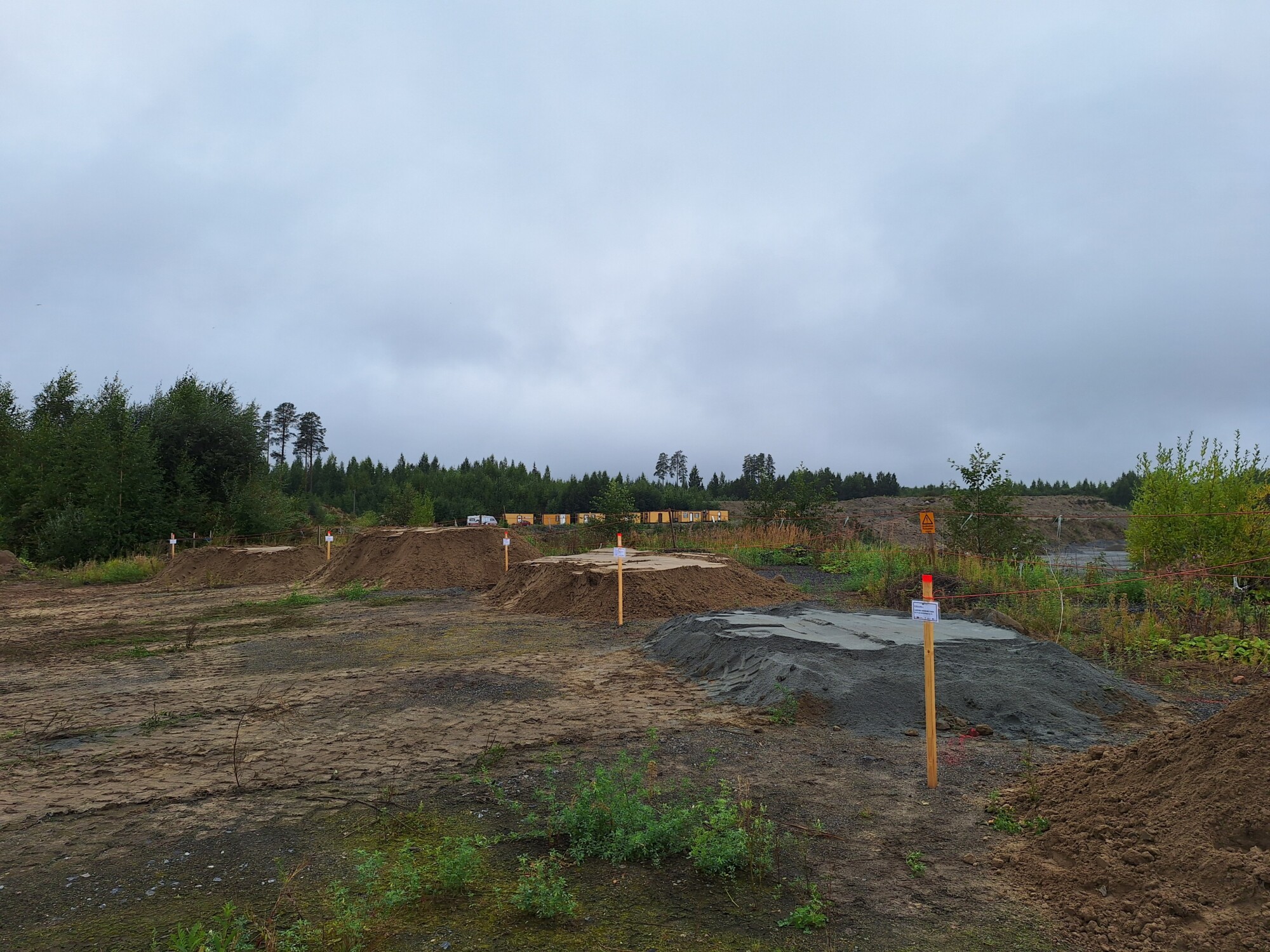 Avoin, hiekkainen työmaa, jossa on useita puisia merkkejä ja pieniä sorakasoja. Puut ja pensaat reunustavat aluetta pilvisen, pilvisen taivaan alla.