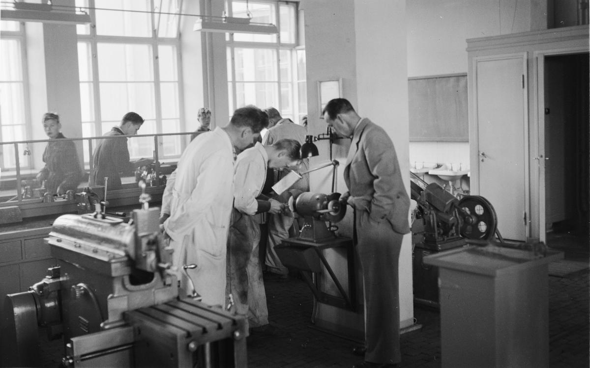 Ryhmä miehiä, joista osa laboratoriotakkisissa, kokoontuu työpajan tai laboratorion koneen ympärille tarkkailemaan tai työskentelemään laitteiden parissa, ja taustalla on suuria ikkunoita, joista pääsee luonnonvaloa sisään.