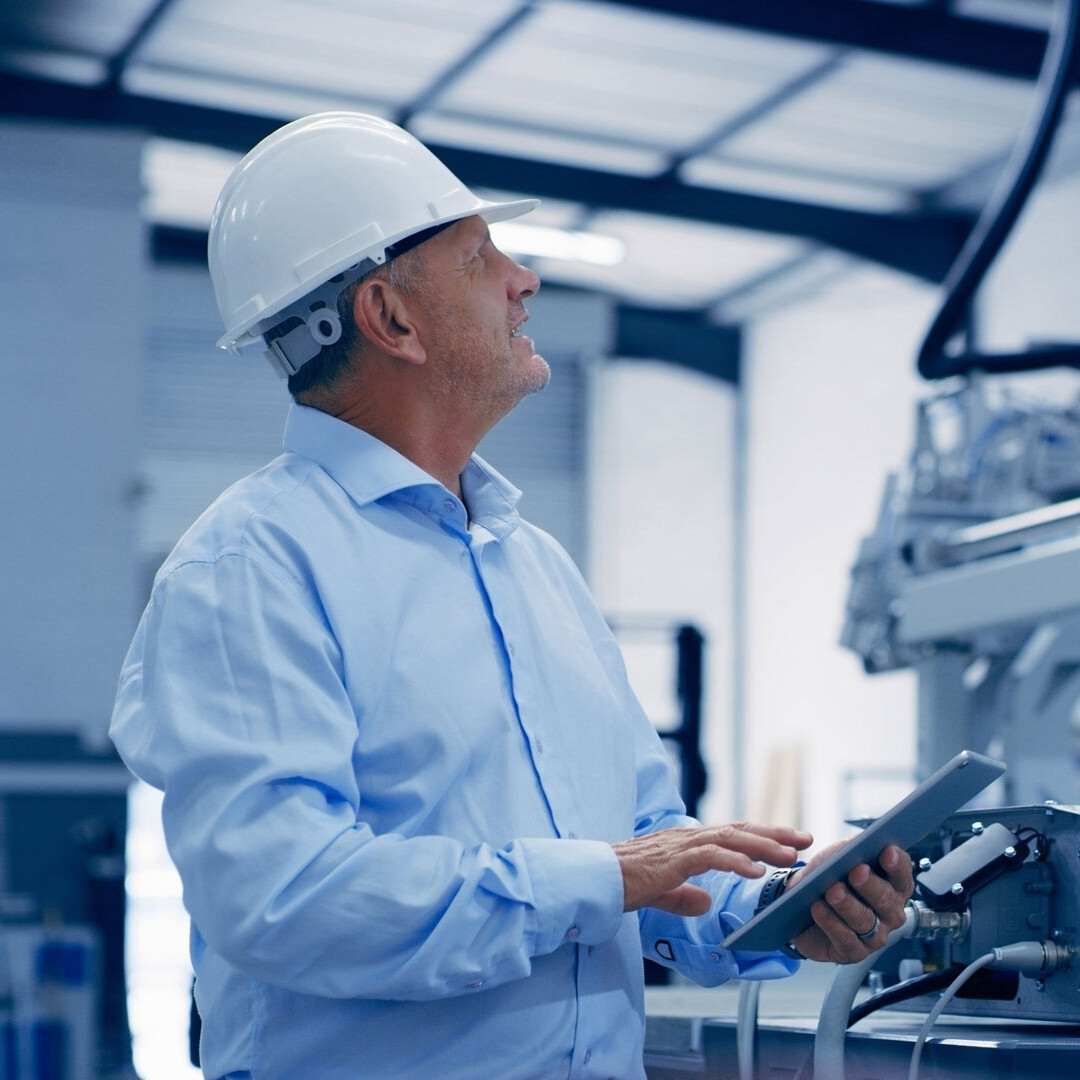 Insinööri, jolla on valkoinen suojahattu ja sininen paita, tarkastaa teollisuuskoneita pitäen kädessään tablettia modernissa tehdasympäristössä.