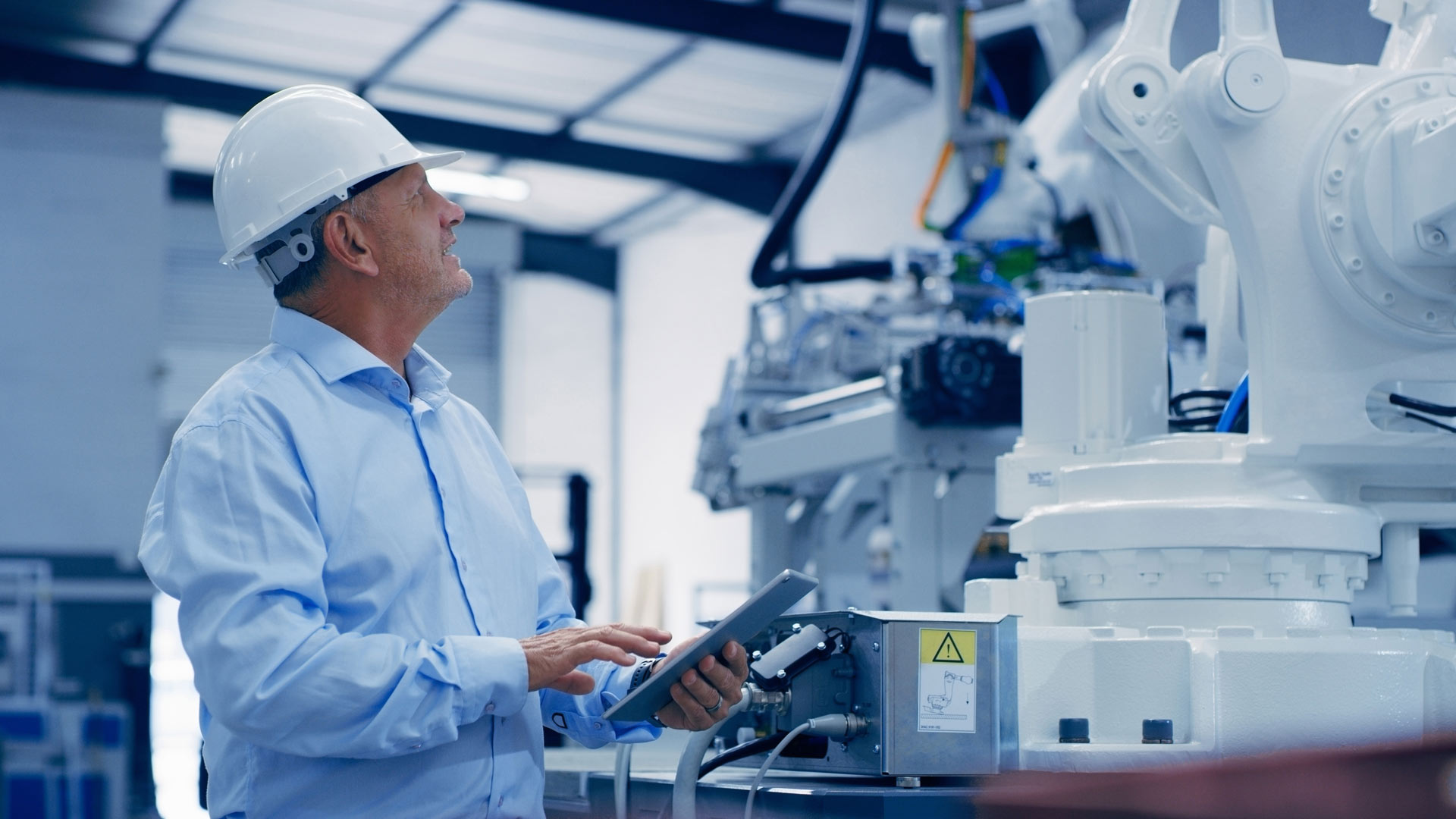 Insinööri, jolla on valkoinen suojahattu ja sininen paita, tarkastaa teollisuuskoneita pitäen kädessään tablettia modernissa tehdasympäristössä.
