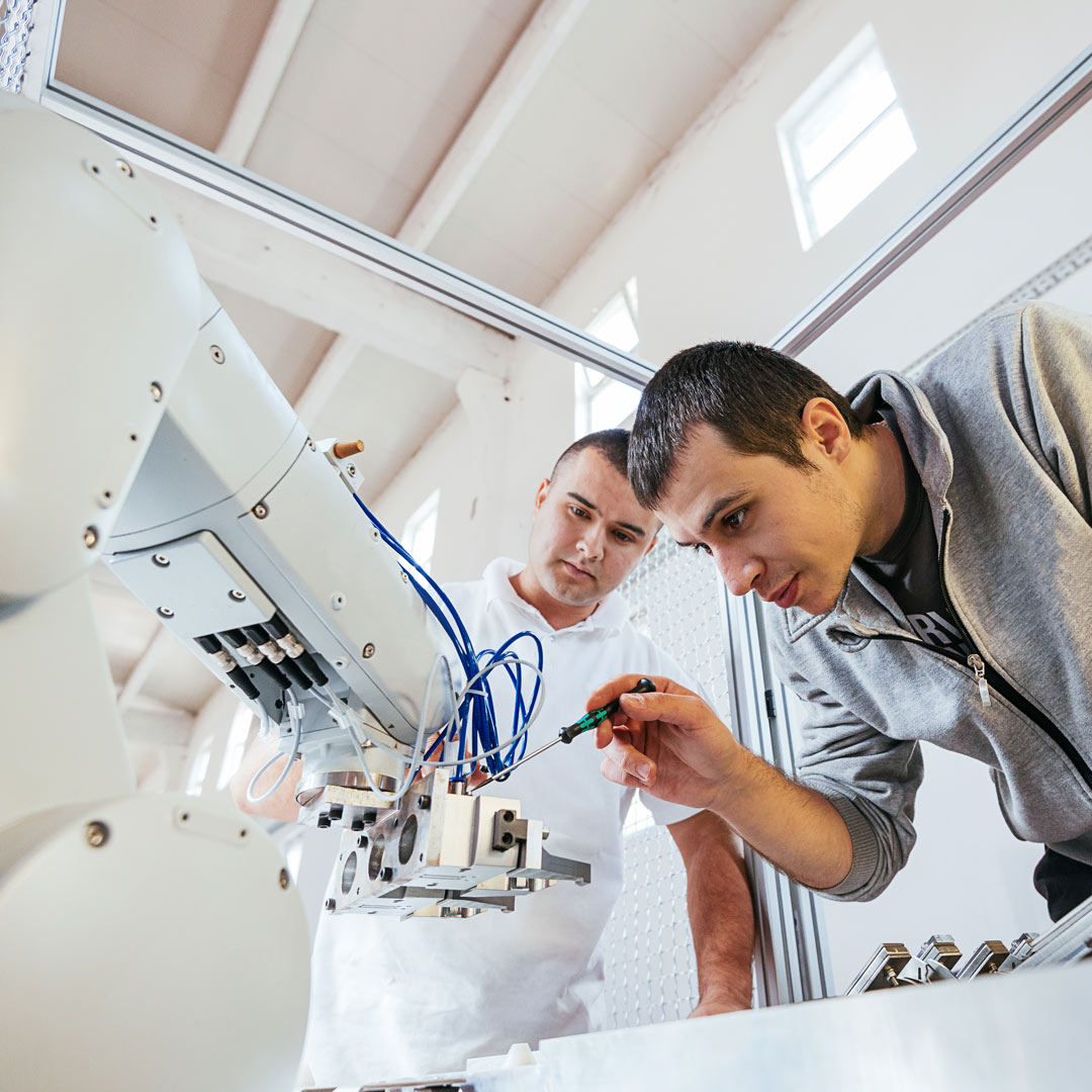 Kaksi miestä tutkii ja säätää tarkasti teollisen robottikäsivarren osia valoisassa, modernissa laboratorioympäristössä.