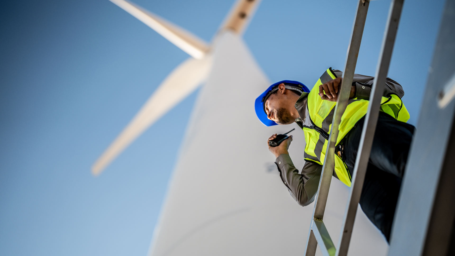 Keltaiseen turvaliiviin ja siniseen suojakypärään pukeutunut työntekijä seisoo metallisella korokkeella ja puhuu radiopuhelimeen, ja taustalla on suuri tuuliturbiini ja kirkkaan sininen taivas.