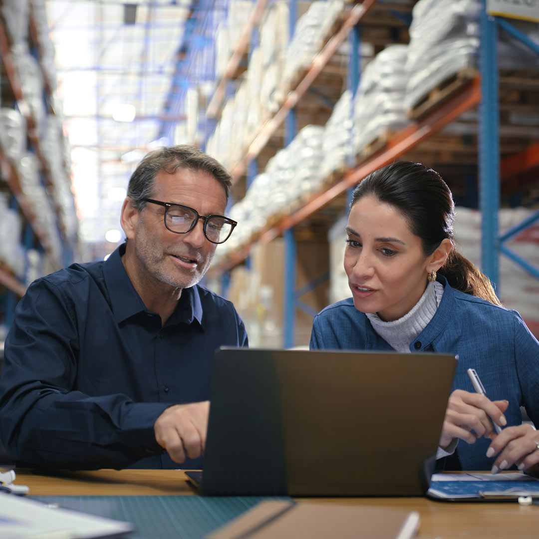 Kaksi ihmistä istuu työpöydän ääressä varastossa ja katsoo yhdessä kannettavan tietokoneen näyttöä. Taustalla on paketteja täynnä olevia hyllyjä. Molemmat vaikuttavat keskittyneiltä ja sitoutuneilta työhönsä.