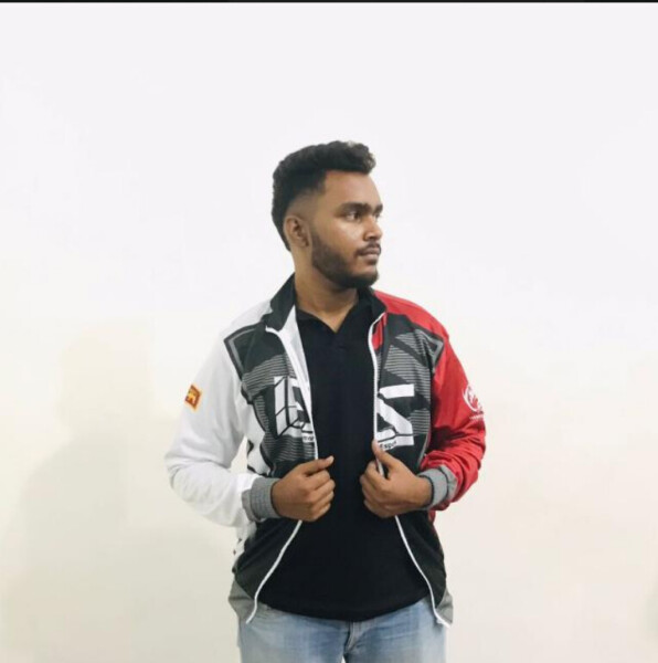 Lyhythiuksinen, parrakas mies seisoo valkoista seinää vasten ja katsoo vasemmalle. Hänellä on yllään musta paita, vaaleansiniset farkut ja vetoketjullinen takki, jossa on punaisia, valkoisia ja mustia geometrisia kuvioita.