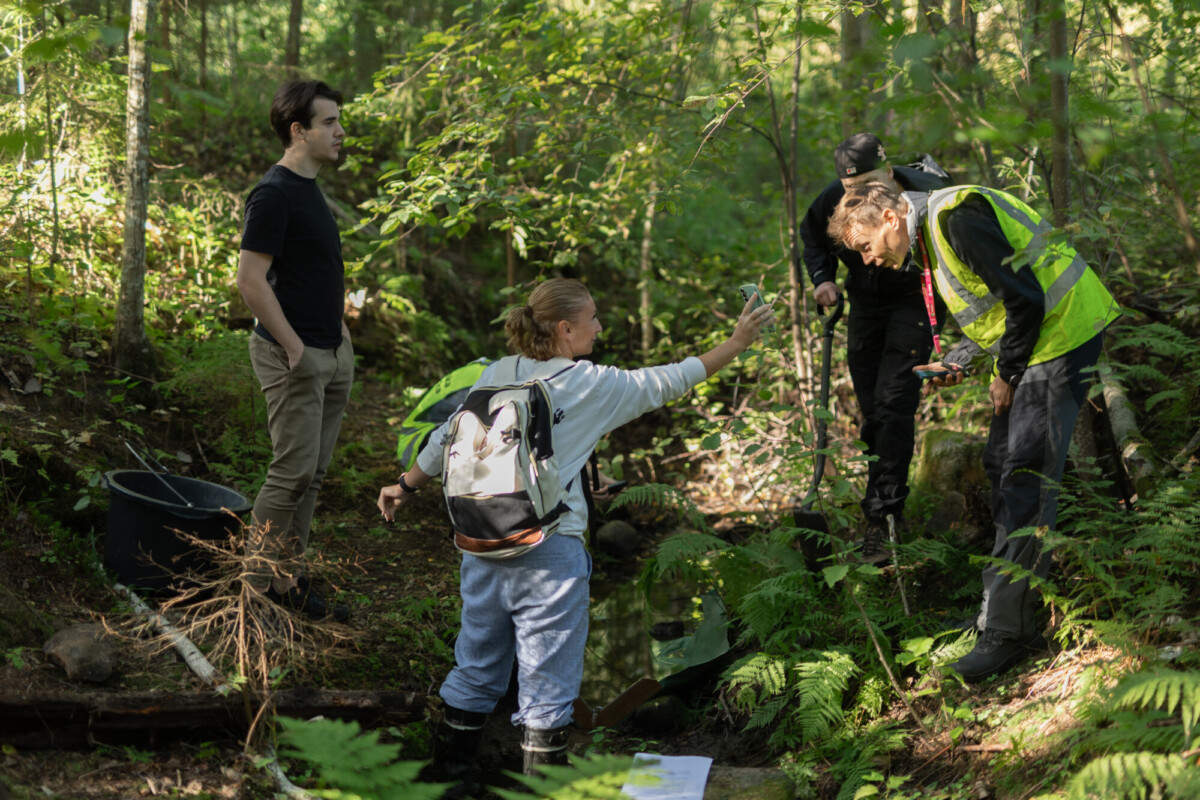 Neljä ihmistä seisoo aurinkoisessa, vihreässä metsässä pienen puron lähellä. Yksi näkösuojaliiviin pukeutunut henkilö tutkii jotakin maassa, kun taas toinen henkilö viittoo heitä kohti. Ryhmä näyttää tekevän kenttätyötä tai tutkimusta.