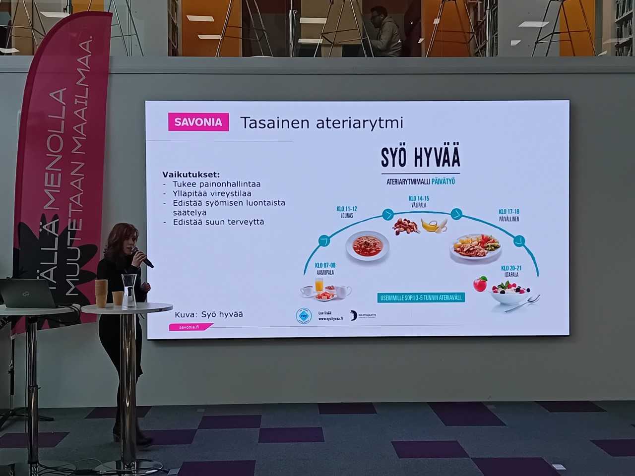 Nainen esittelee suuren näytön edessä suomalaista infograafia tasapainoisesta ateriarytmistä, jossa on ateria-aikoja, terveellisiä ruokakuvia ja luettelo hyödyistä, kuten painonhallinnasta ja hyvinvoinnista. Vasemmalla näkyy vaaleanpunainen Savonia-banneri.
