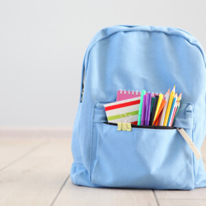 Puulattialla on vaaleansininen reppu, jonka etutaskussa on värillisiä kyniä, vihkoja ja kansioita, ja sen taustana on vaalea seinä.