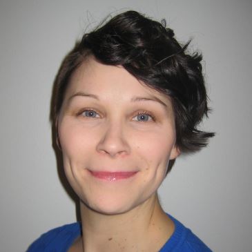 Henkilö, jolla on lyhyet, tummat, aaltoilevat hiukset, hymyilee kameralle. Hänellä on yllään sininen paita ja hän seisoo tavallisen, vaalean taustan edessä.