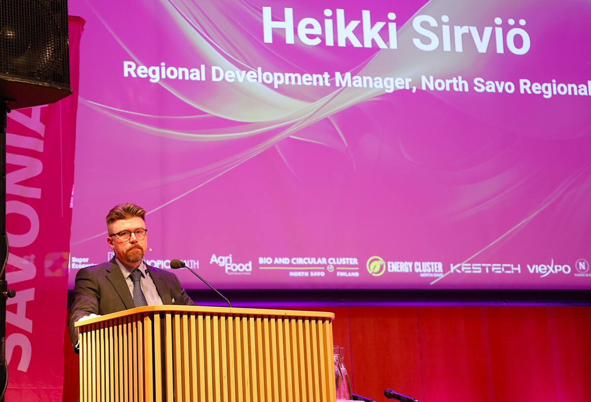 Silmälasipäinen, parrakas mies seisoo puhujakorokkeella, ja hänen takanaan suurella näytöllä näkyy Pohjois-Savon aluekehityspäällikkö Heikki Sirviö ja useita logoja kirkkaan vaaleanpunaisella pohjalla.