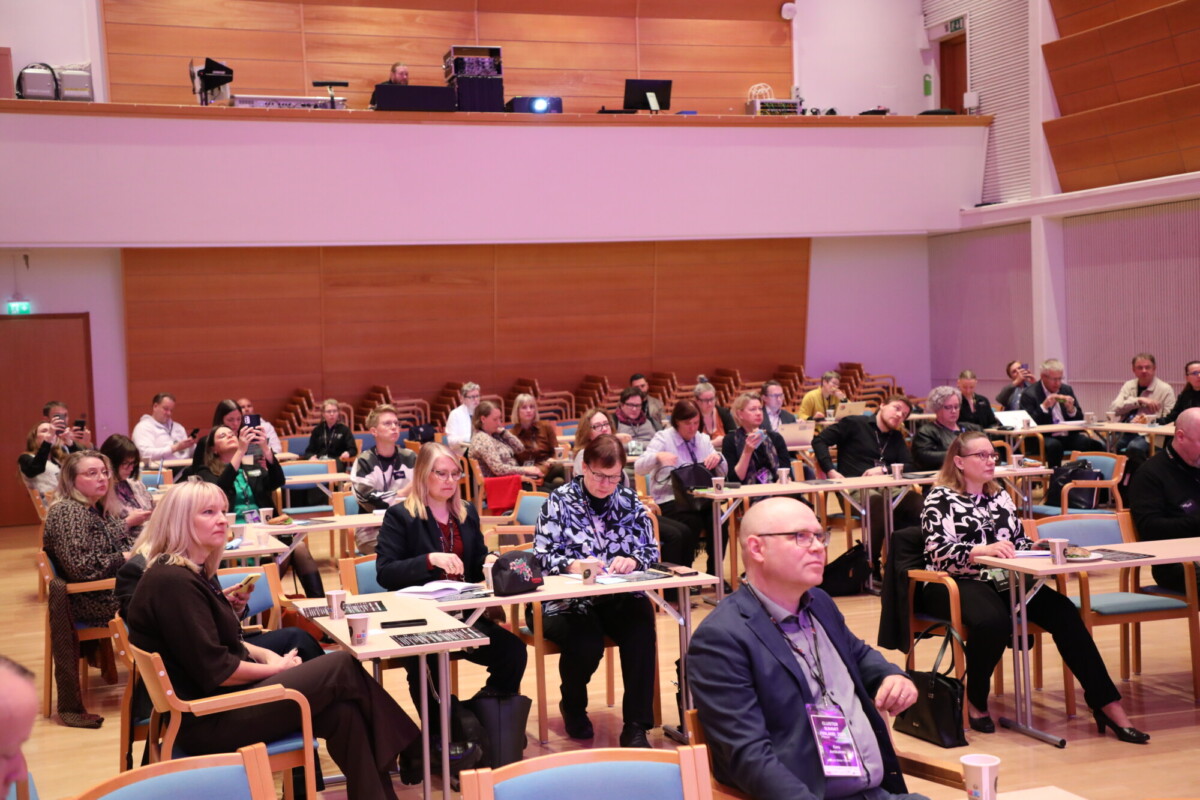 Joukko ihmisiä istuu neuvotteluhuoneen pöydissä ja kuuntelee tarkkaavaisesti esitystä. Huoneessa on puiset seinät ja tuolirivit, ja osallistujilla on pöydillä muistikirjoja ja kannettavia tietokoneita.